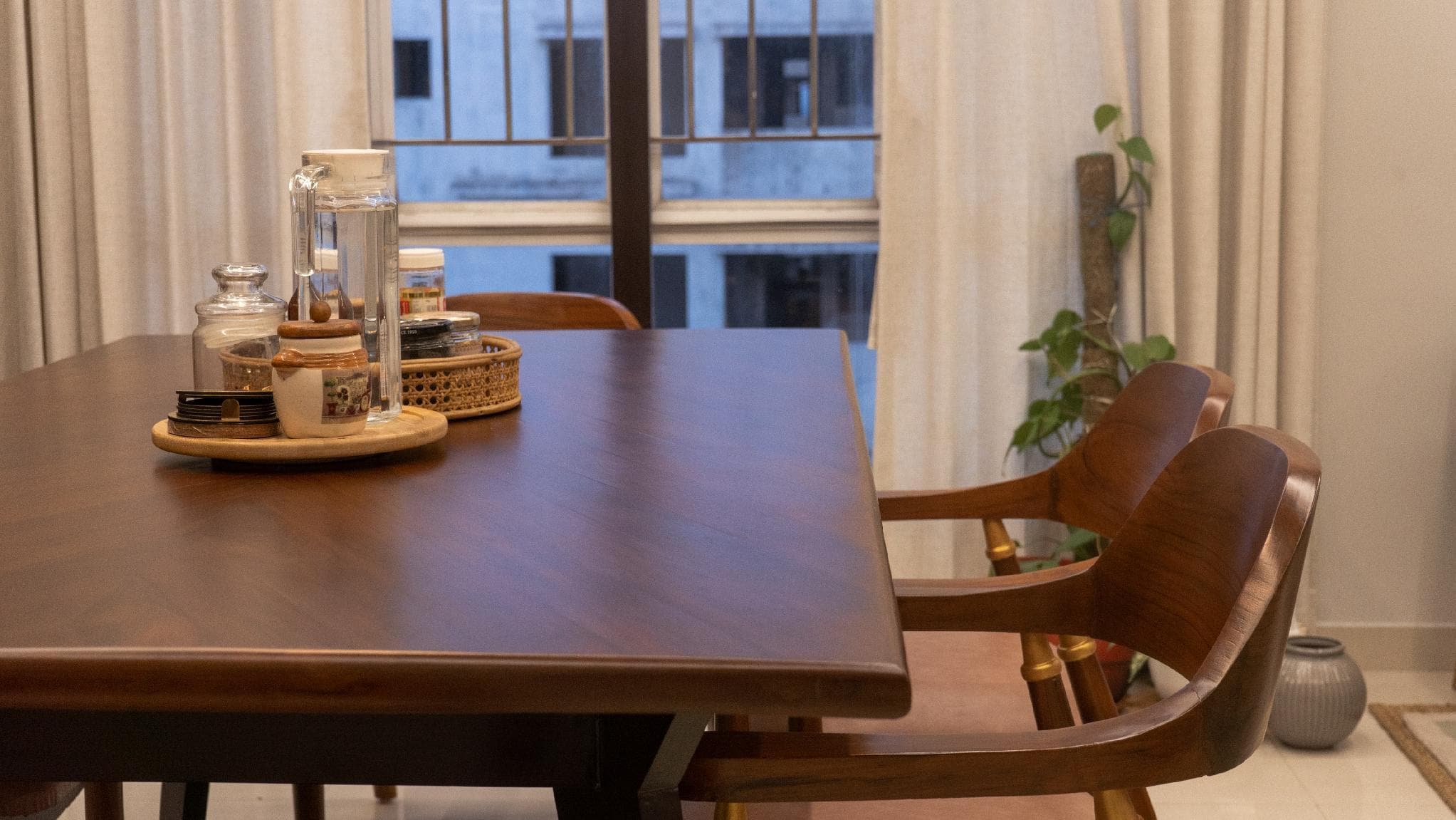 A warm wood dining table set with glass jars and linen curtains beside a window.